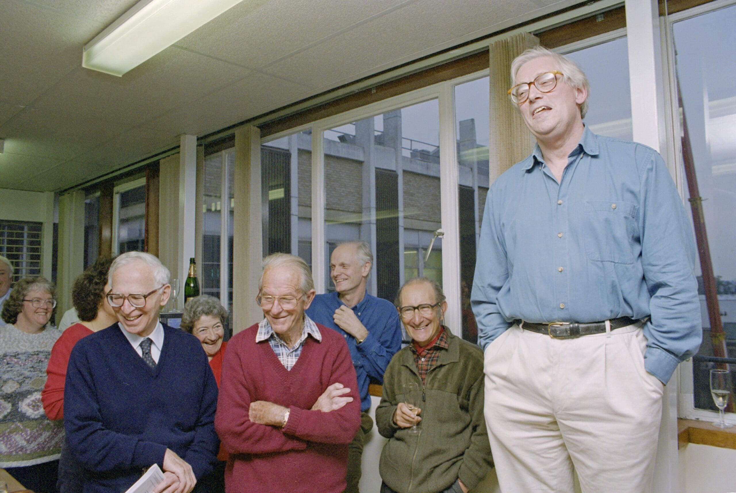 John Walker stood on a chair making a speech. Beside him are stood Aaron Klug, Fred Sanger and César Milstein