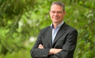 A man stood with his arms crossed in front of a leafy, blurred background