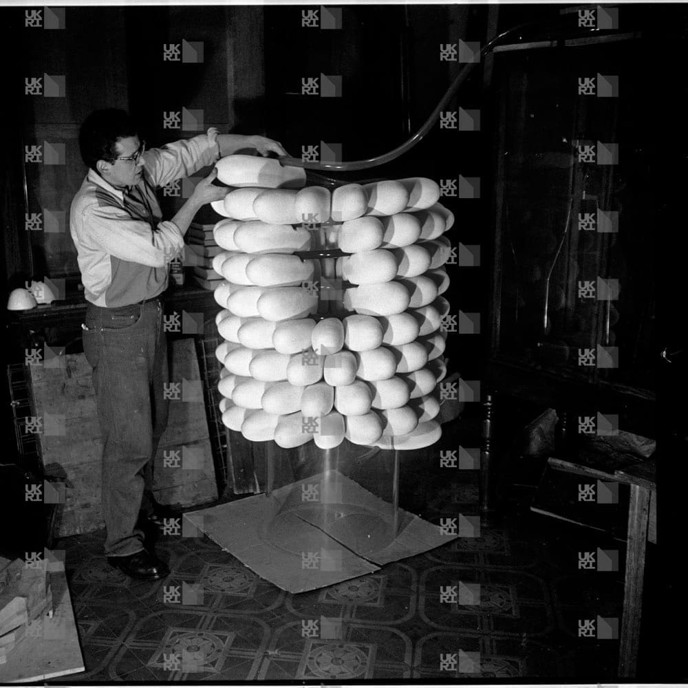 Man in glasses builds a tall, cylindrical sculpture made of stacked rounded objects in a dimly lit room with LMB logo watermark overlay.