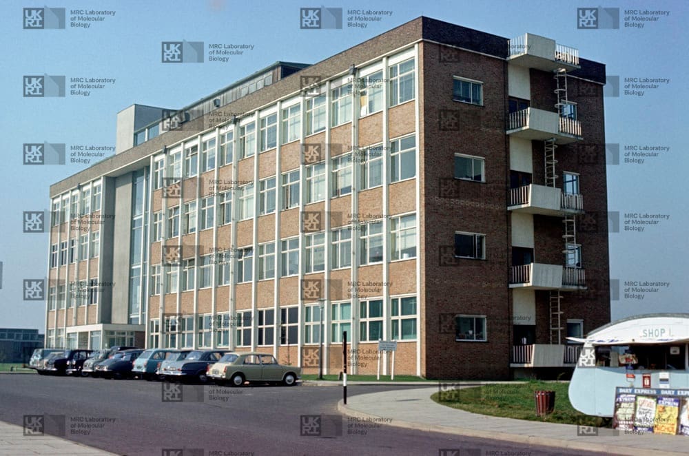1960s brick building with many windows, an exterior staircase, a row of vintage cars, and a small shop trailer in front with LMB logo watermark overlay.
