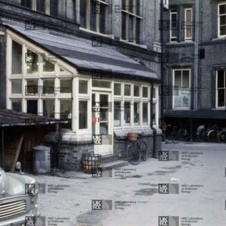 Courtyard with a grey car, a glass-paneled greenhouse structure, and a covered bicycle/scooter parking area in front of a brick building with LMB logo watermark