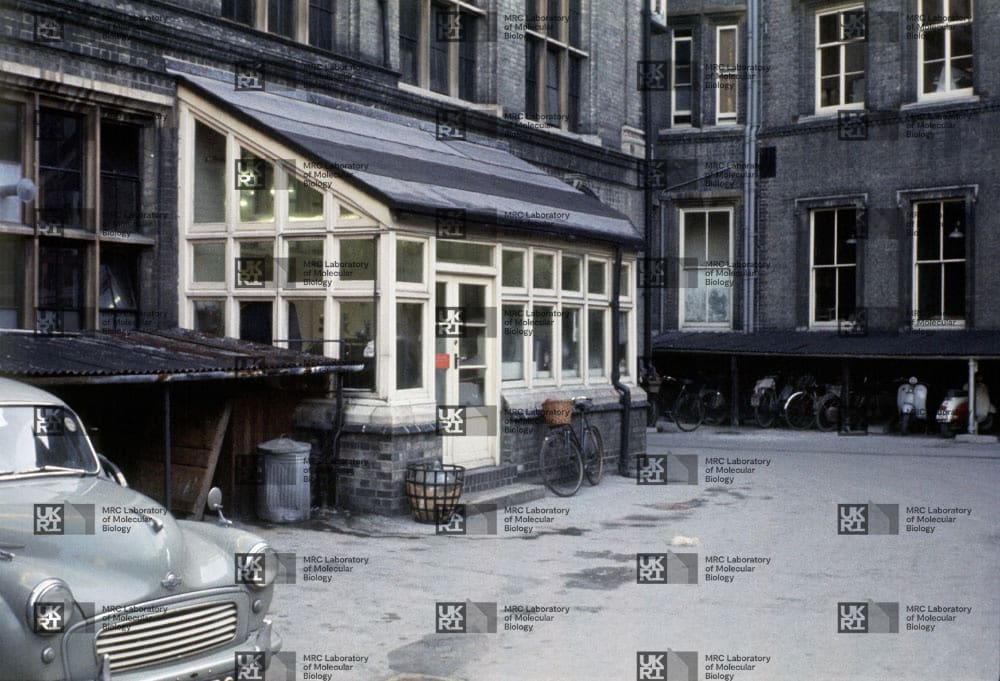 Courtyard with a grey car, a glass-paneled greenhouse structure, and a covered bicycle/scooter parking area in front of a brick building with LMB logo watermark