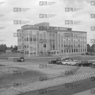 Modern three-story building with many windows, surrounded by a paved lot with several 1960s-era cars, and green areas under an overcast sky with LMB logo watermark