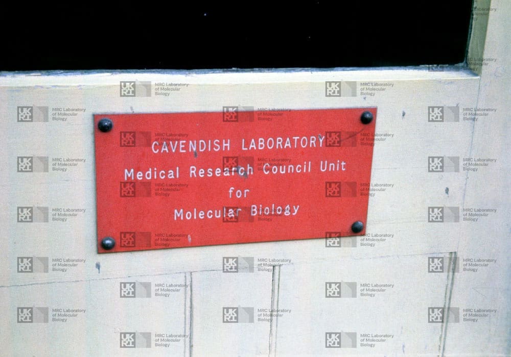 A red rectangular sign for "Cavendish Laboratory Medical Research Council Unit for Molecular Biology" is screwed to a light-colored wall with LMB logo watermark