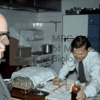 Two men in a kitchen, one in profile, the other smiling as he handles a bottle and glasses on a counter with LMB logo watermark overlayed.