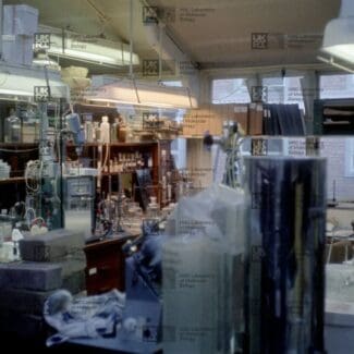 Cluttered laboratory interior filled with scientific equipment, glassware, bottles, shelves, and large cylinders under bright lights with LMB logo watermark