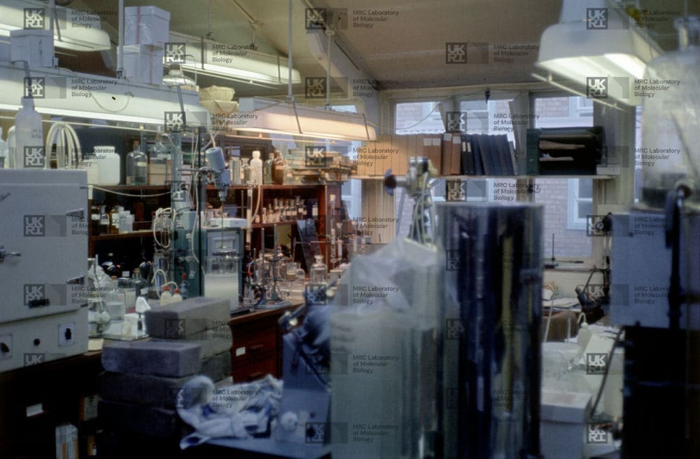 Cluttered laboratory interior filled with scientific equipment, glassware, bottles, shelves, and large cylinders under bright lights with LMB logo watermark