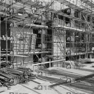 Building under construction covered in scaffolding with piles of pipes and rebar on the ground with LMB logo watermark overlay.