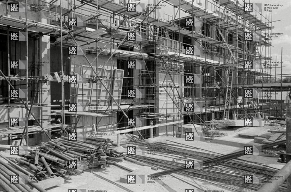 Building under construction covered in scaffolding with piles of pipes and rebar on the ground with LMB logo watermark overlay.