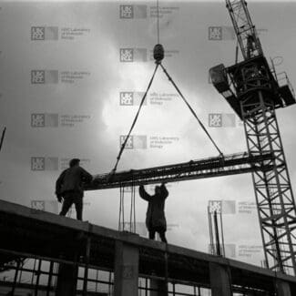 Two construction workers on a building frame guide a long metal beam being lowered by a large crane against a cloudy sky with LMB logo watermark