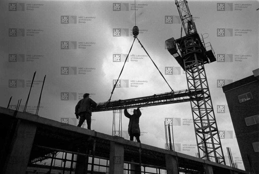 Two construction workers on a building frame guide a long metal beam being lowered by a large crane against a cloudy sky with LMB logo watermark