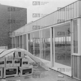 New building extension with many windows and a large stack of metal acoustic tile boxes in the foreground, next to a brick building with LMB logo watermark overlay.