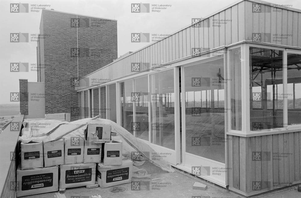 New building extension with many windows and a large stack of metal acoustic tile boxes in the foreground, next to a brick building with LMB logo watermark overlay.