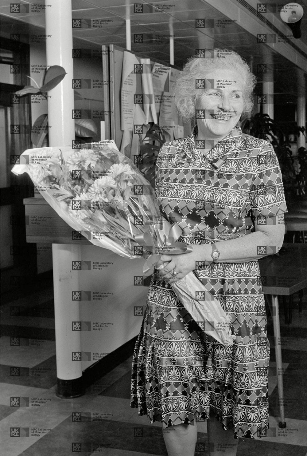 Gisela Perutz holding bouquet of flowers with LMB logo watermark overlay.