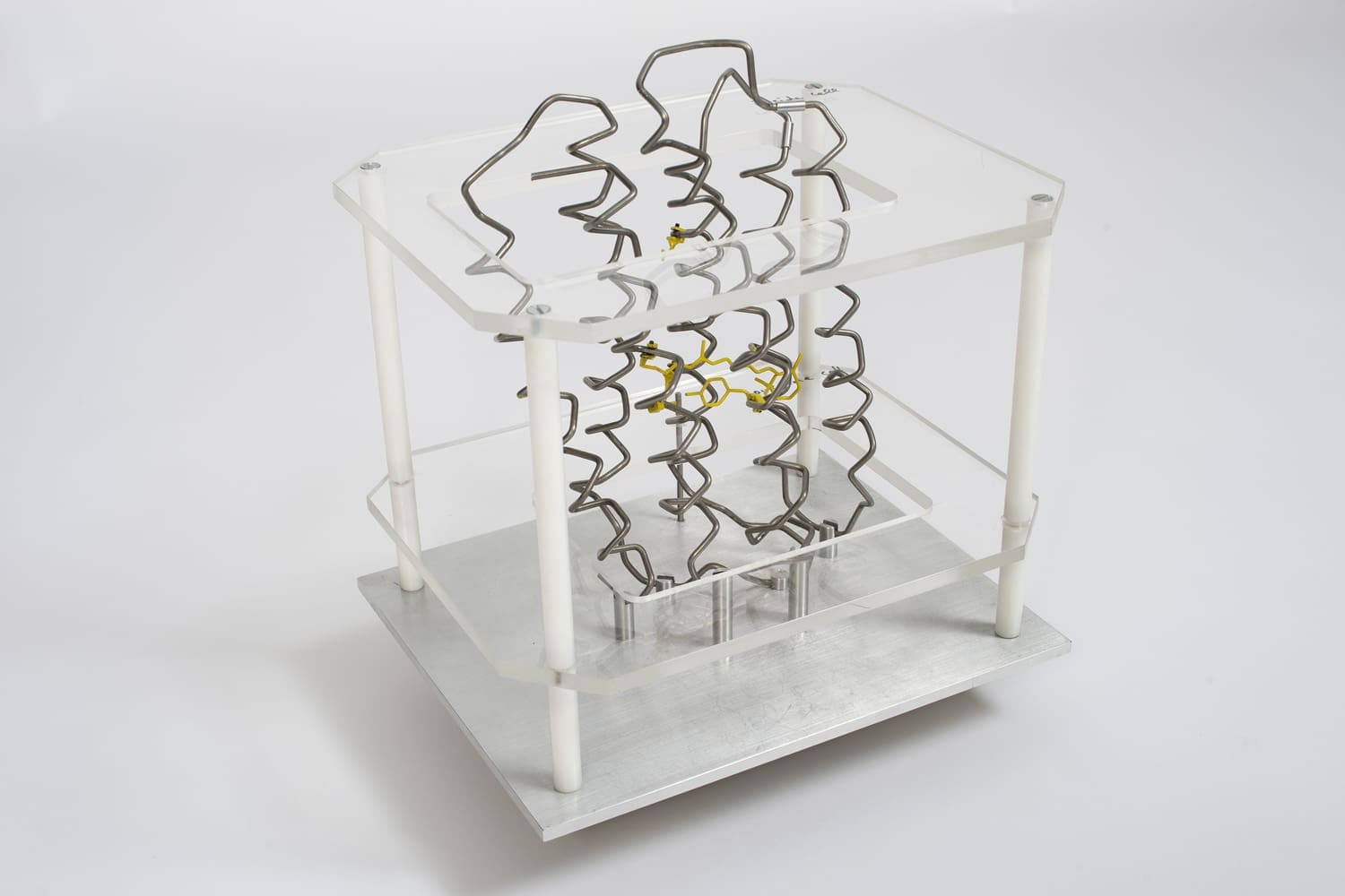 Box structure with transparent plastic bases, white rods, and a metal base, housing spiralled silver metal rods and some smaller yellow metal roads