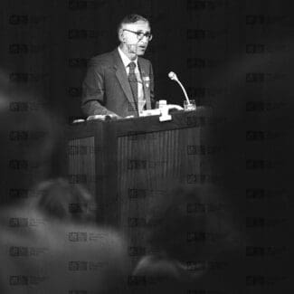 Aaron Klug speaking at lecturn with LMB logo watermark overlay.