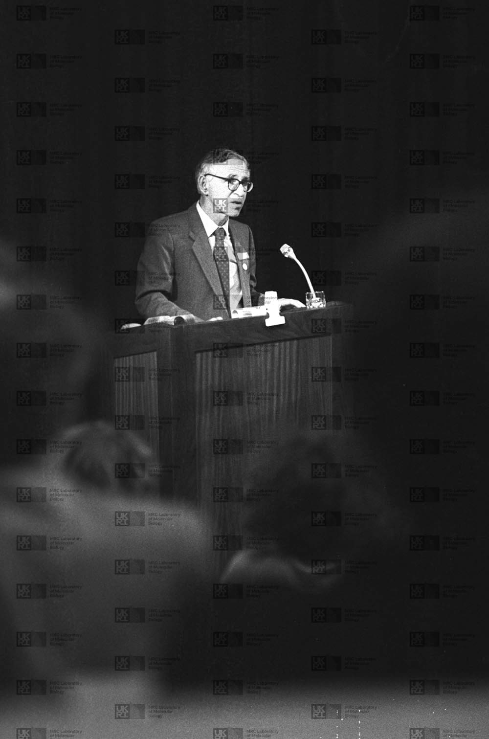 Aaron Klug speaking at lecturn with LMB logo watermark overlay.