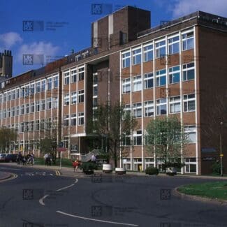 Large brick building with many windows, a spiral fire escape, and a tall chimney structure, next to a curving road with cars and pedestrians with LMB logo watermark overlay.