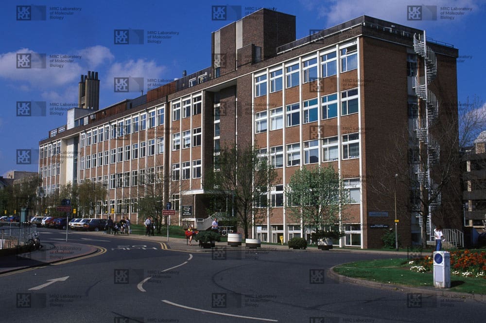 Large brick building with many windows, a spiral fire escape, and a tall chimney structure, next to a curving road with cars and pedestrians with LMB logo watermark overlay.