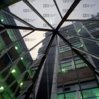 View up through a glass pyramid skylight, revealing parts of a multi-story brick building with green-tinted windows with LMB logo watermark overlay.