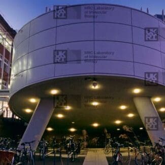 Cylindrical building on stilts, illuminated underneath, surrounded by multi-story brick buildings and many parked bicycles at night with LMB logo watermark overlayed.