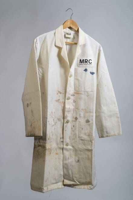 White lab coat with ‘LMB’ written over the pocket, and various chemical stains, on a wooden hanger