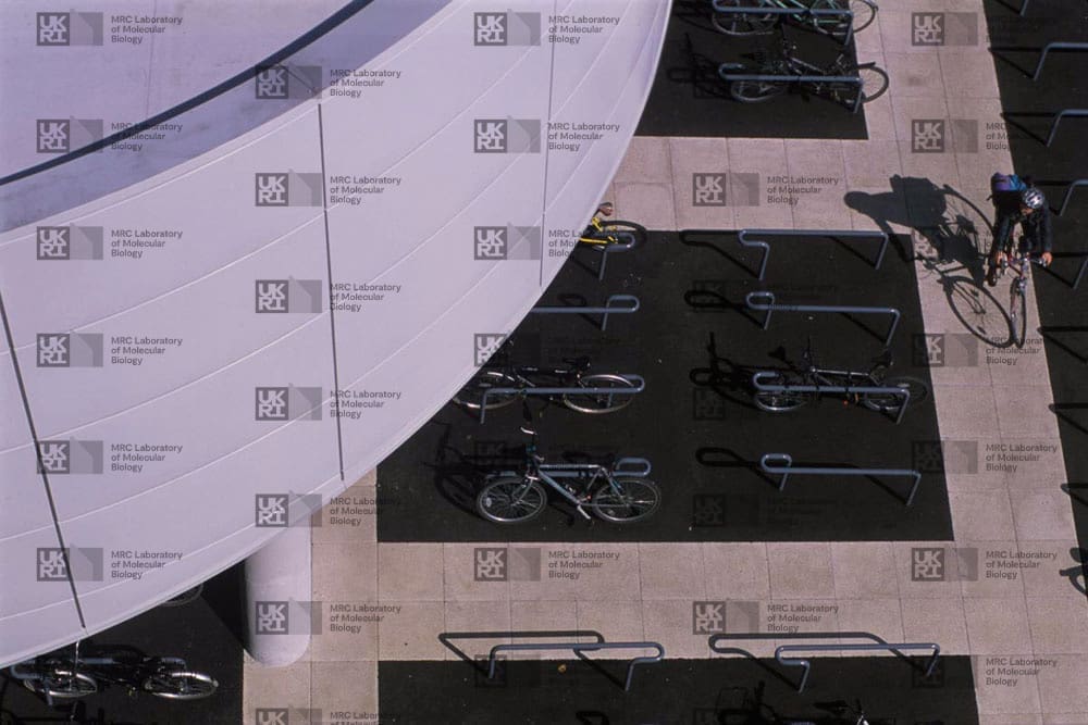 High-angle view of a modern building's curved white facade and an adjacent outdoor area with bicycle racks, parked bikes, and a cyclist, all covered by faint institutional watermarks.