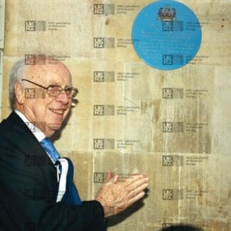 James Watson clapping, a blue plaque is on the wall above him, with LMB logo watermark overlay.