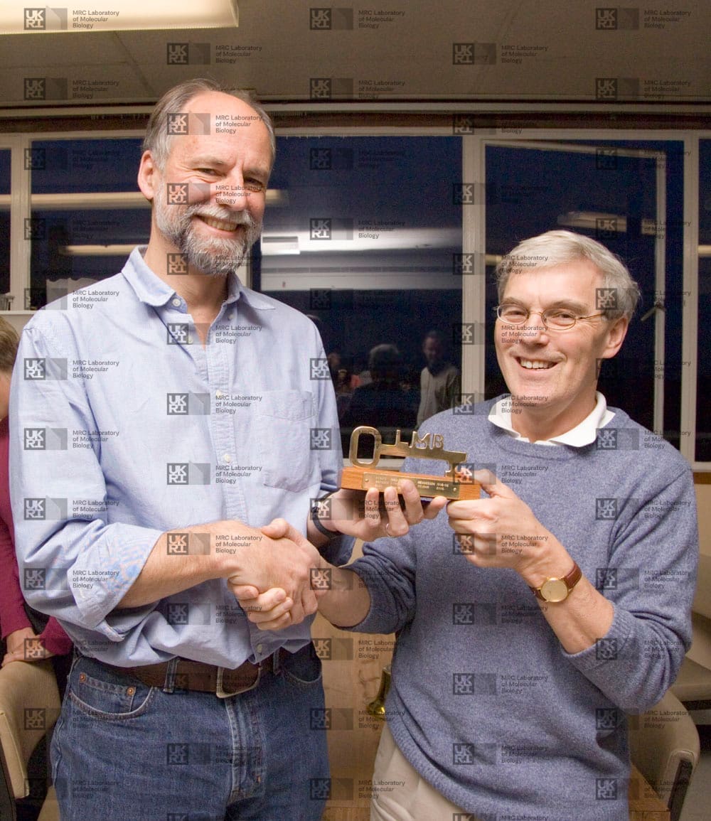 Hugh Pelham and Richard Henderson shaking hands, both holding a key, with LMB logo watermark overlay.
