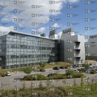 Modern building with glass facade and angular grey sections, surrounded by a parking lot with cars and green landscaping under a partly cloudy sky with LMB logo watermark overlayed.