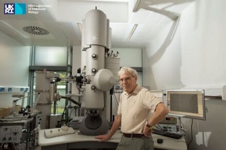 Man stood in front of a large scientific microscope with other surrounding equipment and computer monitors