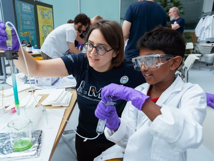 Woman and boy in lab coats and goggles conduct a science experiment with tubes and green liquid, both smiling.