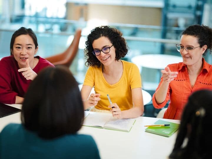 Three women sit at a table smiling and gesturing during a discussion, with one woman's back to the viewer and notebooks on the table.