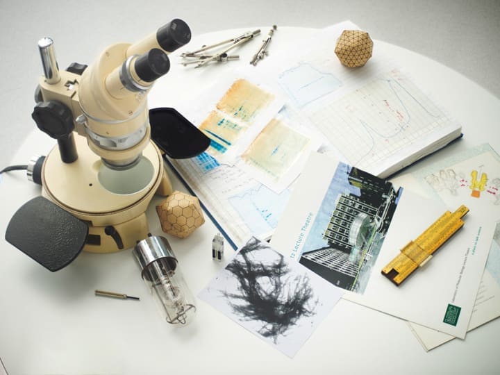 A cream microscope, scattered papers with graphs and images, a vacuum tube, and various scientific tools on a white table.