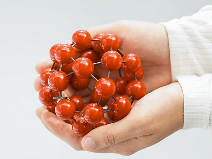 Hands cup a spherical model made of glossy red balls connected by metal rods against a light background.