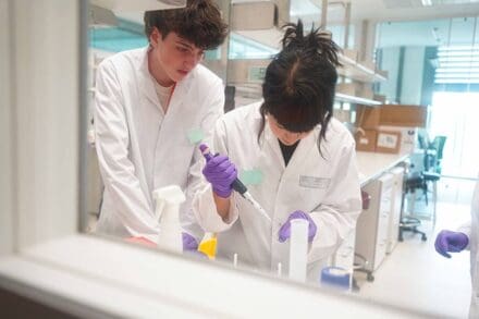 Two students in white lab coats, one operating a pipette into a container with purple gloves, while the other observes in a laboratory.