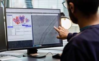 Person in glasses points at a computer monitor displaying scientific data, including colorful molecular models and a grayscale image.