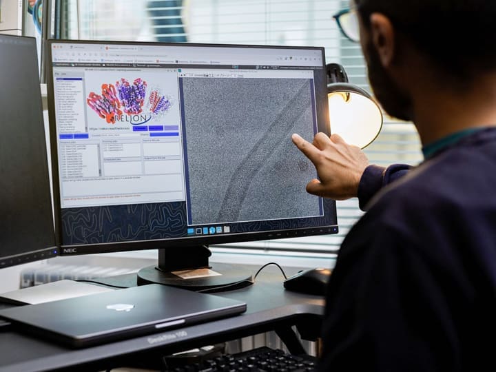 Person from behind points at a computer screen displaying molecular structures and microscopy data in RELION software. A laptop rests on the desk.
