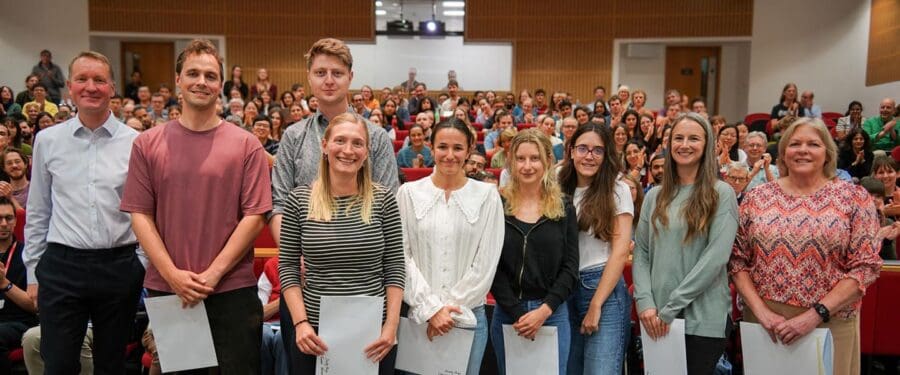 Group of eight prize winners standing on a stage with an audience