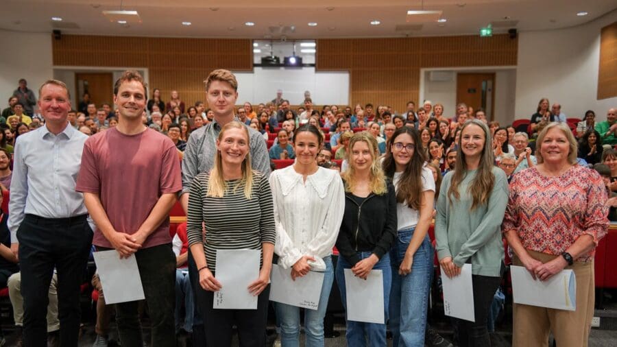 Group of people holding certificates, stood in front on packed lecture theatre