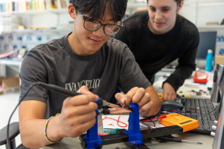 Young man in glasses soldering a circuit board with a woman looking over his shoulder, surrounded by electronic equipment on a workbench.