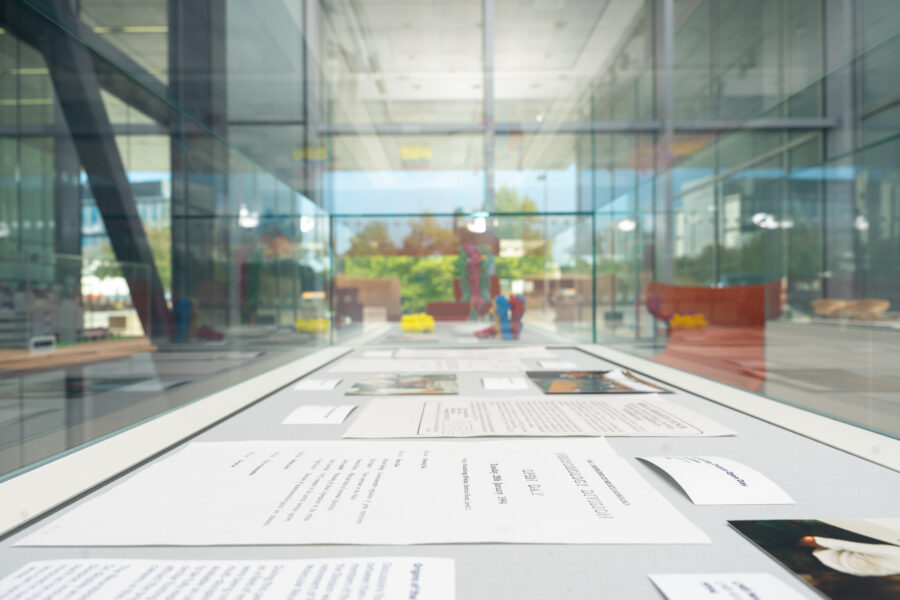 Documents and photographs are displayed in a glass case, with a blurred modern building and trees visible through large windows in the background.