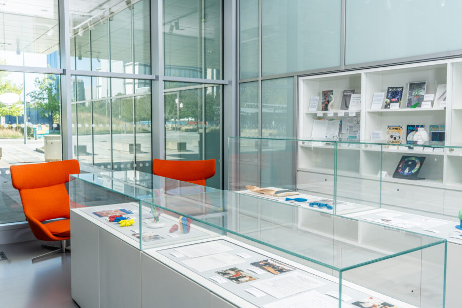 Modern exhibition space with orange chairs, glass display cases holding papers and colorful objects, and wall shelves with journals, next to large windows.