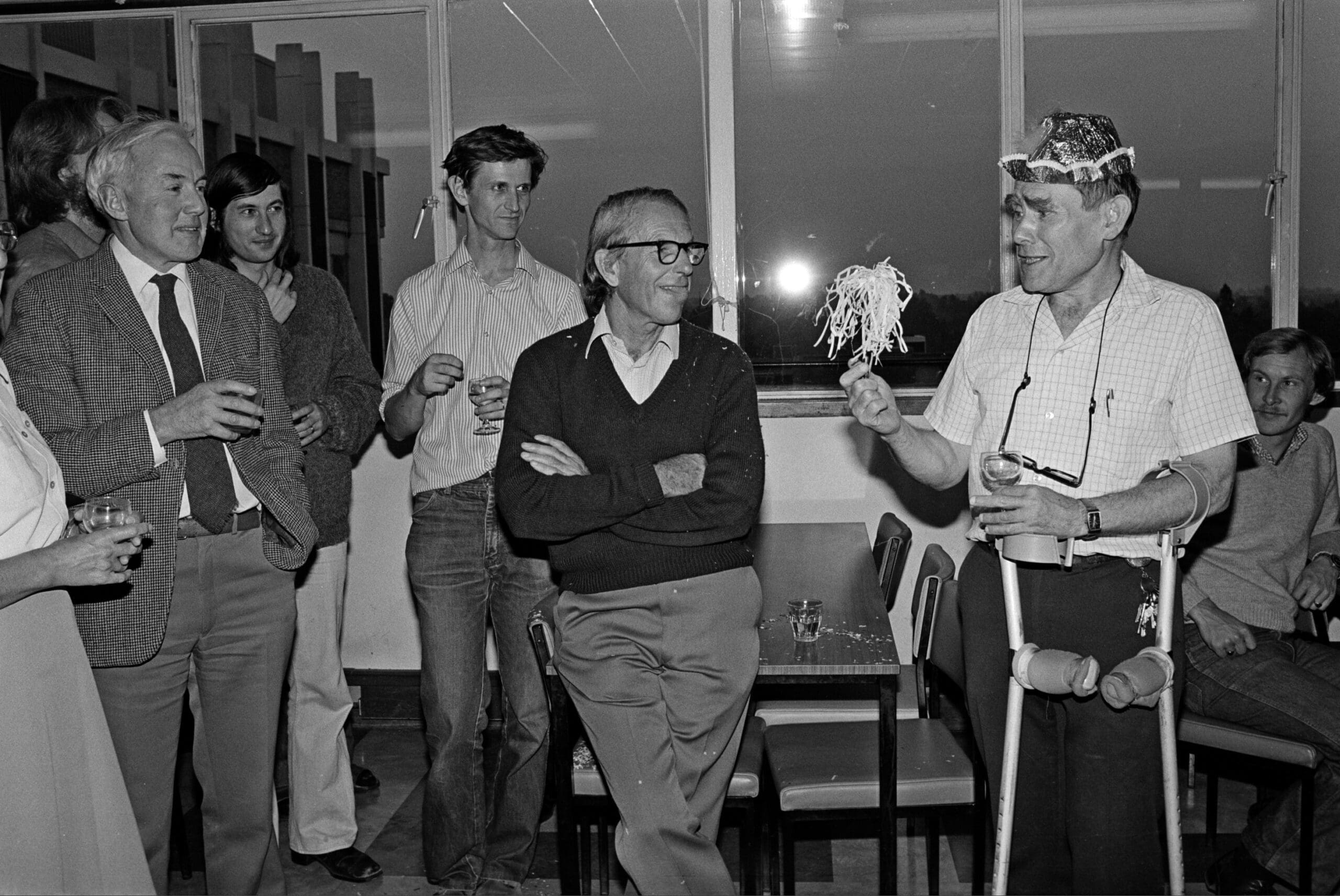 Fred Sanger, centre, looking at Sydney Brenner, right, who is wearing a party hat and holding a party streamer. To the left, people stand holding wine glasses