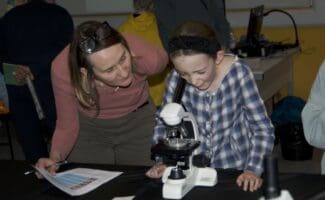 Woman and girl intently observe a white microscope, the girl peering into its eyepiece while the woman holds a paper.