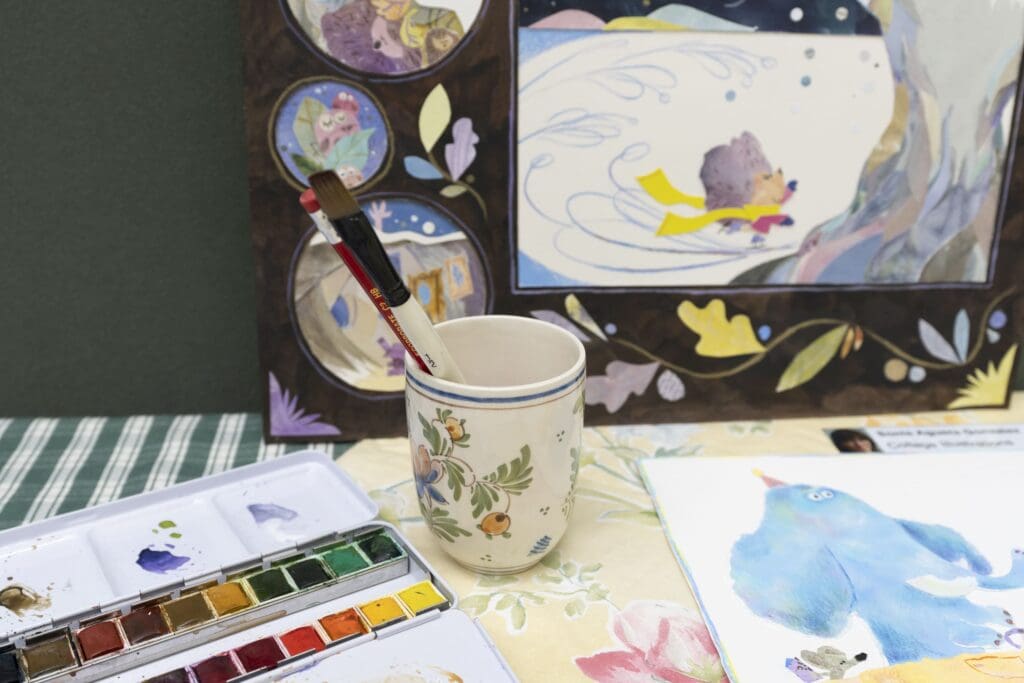 Ceramic cup holding a brush and pencil , and surrounded by a watercolour paint palette, and illustrations depicting animal characters.