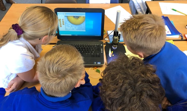 Four children examine a laptop displaying a magnified image from a digital microscope on a wooden desk.