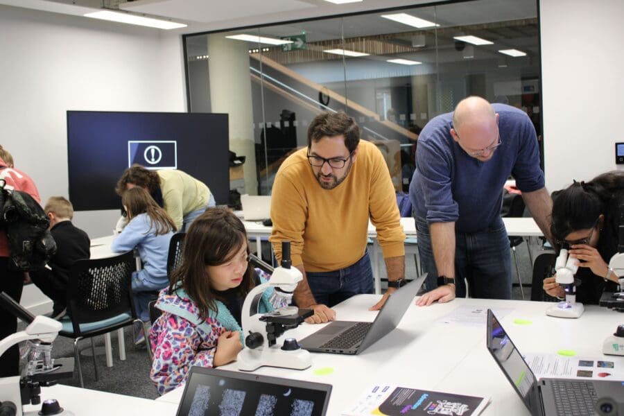 Children and adults engage with microscopes and laptops at white tables in a bright, modern room.
