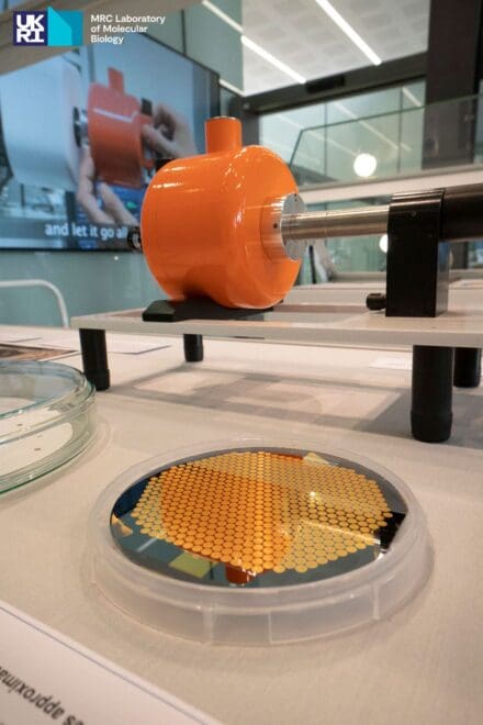 Cylindrical plate with gold honeycomb design in the foreground, with orange scientific equipment in the background