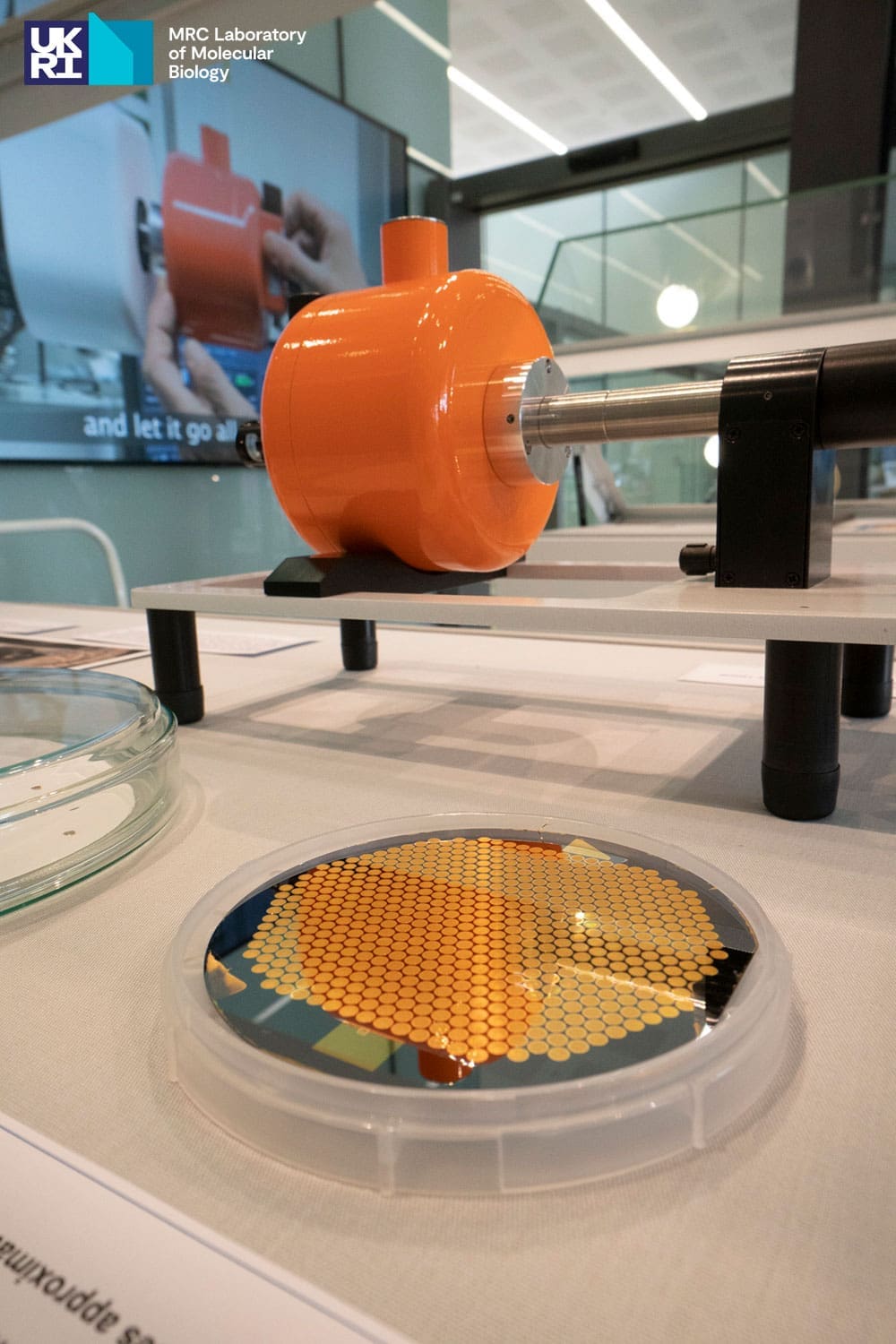 Cylindrical plate with gold honeycomb design in the foreground, with orange scientific equipment in the background